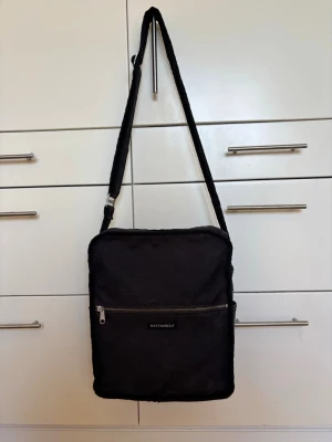 Svart axelväska från Marimekko 29X34X12 cm, rymmer laptop o A4 - Snygg svart axelväska från Marimekko i slitstark canvas. Väskan har justerbar axelrem, en stor ficka med dragkedja framtill och en öppen ficka på baksidan. Perfekt för dig som vill ha en stilren och praktisk väska till vardagen.