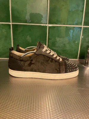 Christian Louboutin  - Unika camo sneakers med mockaliknande material och vita sulor. Skorna har vita snören och coola silverfärgade nitar på tån som ger en edgy look. Perfekta för dig som vill sticka ut och addera lite attityd till din outfit.
