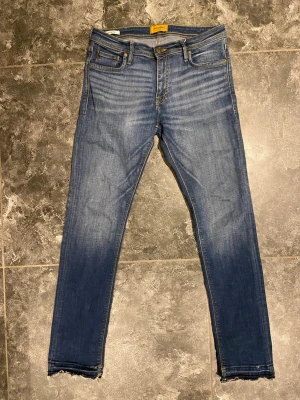 Blå jeans med rå kant Jack & Jones - Snygga blå jeans med slitningar och rå kant vid bensluten. Jeansen har klassisk femficksmodell, normalhög midja och ljusa tvättade partier framtill och baktill för en trendig look. Perfekta för dig som gillar en avslappnad men ändå stilren stil.