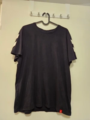 T-shirt från Hummel - Svart t-shirt med korta ärmar och rund halsringning. Tröjan har vita ränder på ärmarna.  Nytvättad med parfymfritt tvättmedel.