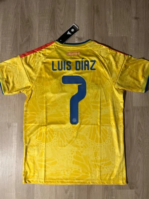 Colombia landslagströja Luis Díaz #7 - Gul Colombia fotbollströja med blå detaljer, röd axelrand och nummer 7. På bröstet finns landslagets emblem och på ryggen står det 'Luis Díaz' samt stort nummer 7. Tillverkad i lätt och ventilerande material. HELT NY!