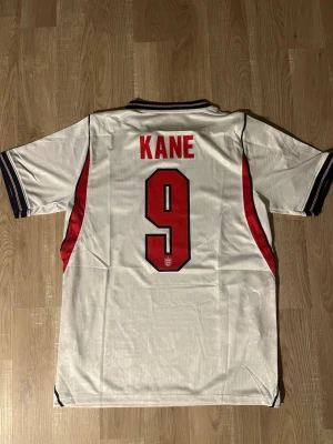 England VM-fotbollströja nr 9 Kane - Vit England fotbollströja med svarta och röda detaljer, nummer 9 och namnet Kane på ryggen. Tröjan har korta ärmar, broderat landslagsmärke på bröstet. Perfekt för fotbollsfans som vill visa sitt stöd. HELT NY!