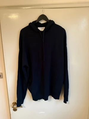 Mörkblå hoodie med huva - En stilren mörkblå hoodie med huva och långa ärmar. Hoodien har en enkel design utan tryck eller mönster och är tillverkad i ett mjukt material som passar perfekt för en avslappnad look. Dragsko i huvan för justerbar passform.
