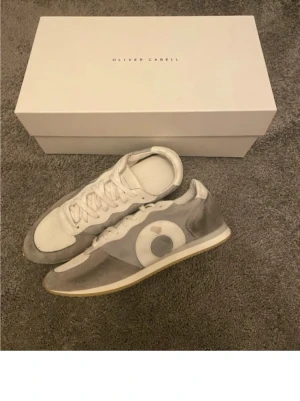 Gråvita sneakers från Oliver Cabell - Snygga sneakers i grått och vitt från Oliver Cabell med klassisk låg siluett. Ovandelen är i mocka och mesh med en vit cirkeldetalj på sidan. Yttersulan är i gummi med beige ton. Perfekta för en clean och stilren look.