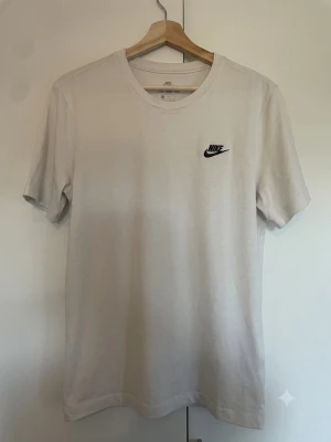 Vit t-shirt från Nike med logga - Klassisk vit t-shirt från Nike med svart logga på bröstet. T-shirten har rund halsringning och korta ärmar. Tillverkad i mjuk bomull för en skön och avslappnad stil. Perfekt till jeans eller shorts. Har en lite fläck som knappt syns (se sista bilden)