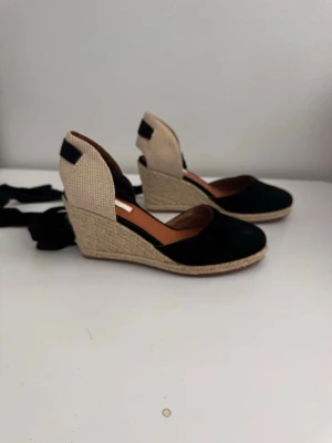 Svarta espadrille ballerinaskor med kilklack - Säljer ett par snygga ballerinaskor i espadrille-stil med svart ovandel i tyg och beige hälparti. Skorna har kilklack i flätad jute och svarta knytband runt ankeln. Perfekta för dig som vill ha en trendig och bekväm sko med sommarkänsla.