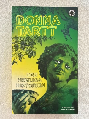 Den hemliga historien av Donna Tartt - En svensk pocketbok med omslag i grönt och gult, skriven av Donna Tartt. Boken har ett konstnärligt omslag med en staty och grafiska detaljer. Romanen är en kultklassiker inom modern skönlitteratur och handlar om mörka hemligheter på ett college. Jag säljer för att jag redan har en💖