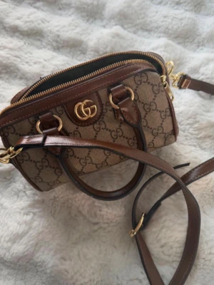  brun  handväska - Säljer en snygg brun handväska från G med klassiskt GG-monogram, guldfärgade detaljer och både handtag och axelrem i mörkbrunt läder. Väskan har dragkedja upptill och är perfekt för dig som gillar lyxig och tidlös design. Köpt för 1200kr och säljs för 800kr.