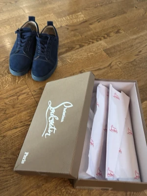 Mörkblå sneakers från Louboutin - Snygga mörkblå sneakers från Christian Louboutin i mocka med klassisk röd sula och svarta snören. Låg modell med rund tå och diskreta sömmar. Perfekta för dig som vill ha något exklusivt och stilrent.men de är lite sönder på höger skon