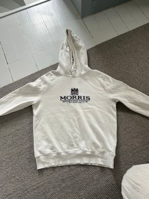 Vit hoodie från Morris Sporting Club - Säljer en vit hoodie från Morris Sporting Club med tryck på bröstet och klassisk huva. Hoodien har ribbade muddar vid ärmslut och nederkant samt en relaxed passform. Perfekt för en chill och stilren look. 