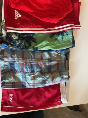 Flerpack badbyxor från Adidas, GANT & BOSS - Säljer ett paket med fyra par badbyxor: ett rött par från Adidas med vita ränder, ett grönt/blått med palmtryck, ett rutigt par från GANT i blå och orange toner samt ett rött par från BOSS med vit logga. Perfekta för stranden eller poolen!