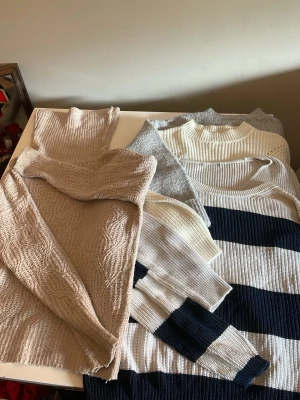 Flera stickade tröjor i neutrala färger - Säljer ett gäng stickade tröjor i beige, grått, vitt och en med breda marinblå och vita ränder. Tröjorna har olika stickade mönster och är långärmade, perfekta för lager-på-lager och mysiga dagar.