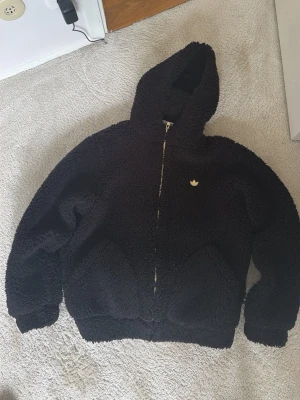 Svart teddyjacka med huva från Adidas - Mysig svart teddyjacka från Adidas med huva och dragkedja framtill. Jackan har ett broderat Adidas-logo på bröstet och är tillverkad i fluffigt teddy-material. Perfekt för kalla dagar när du vill vara både varm och stilren.