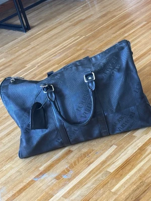 Svart weekendbag - Snygg svart weekendbag ,präglad logotyp över hela väskan. Väskan har dubbla handtag, dragkedja upptill och detaljer i metall. Perfekt för resor och har en klassisk, stilren design i tåligt material.