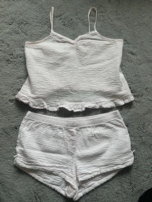 Vitt pyjamasset med linne och shorts - Skriv om du har önska för endast en del! Säljer ett vitt pyjamasset med linne och shorts i mjukt, strukturerat tyg. Linnet har tunna axelband och volangkant nedtill, medan shortsen har söta rosetter på sidan och matchande volangdetaljer. Perfekt för mysiga kvällar hemma eller hos en sleepover.
