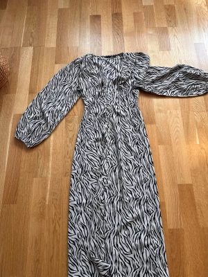 Zebramönstrad oneshoulder klänning - Snygg lång klänning med zebramönster i svart och vitt. Klänningen har oneshoulder-design och en lång puffärm, vilket ger en trendig och unik look. Perfekt för dig som vill sticka ut med ett djurmönster och cool passform.