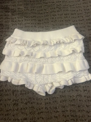 Vita stickade shorts med volanger - Supersöta vita stickade shorts med flera lager volanger och ribbad midja. Shortsen har ett detaljerat stickat mönster och känns både lekfulla och trendiga. Perfekta för varma dagar när du vill ha något bekvämt men ändå snyggt.