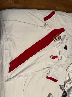 Vit och röd piké från Polo Ralph Lauren - Säljer en vit pikétröja från Polo Ralph Lauren med röd diagonal rand, stor röd logga och broderat klubbmärke på bröstet. Klassisk krage och knappar framtill. Röd detalj på ärmslut och krage. Perfekt för en sportig och stilren look. Storlek M