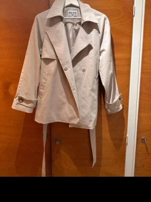 Beige trenchcoat från NA-KD - Stilren beige trenchcoat från NA-KD med klassisk dubbelknäppning, bred krage och bälte i midjan. Jackan har långa ärmar med knappar vid ärmsluten och två snedställda fickor framtill. Perfekt för vår och höst när du vill ha en tidlös look.