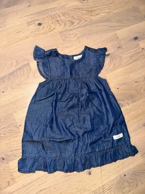 Blå jeansklänning från Newbie - Supersöt blå jeansklänning från Newbie i storlek 80. Klänningen har volangdetaljer vid ärmar och nederkant samt en lös och bekväm passform. Perfekt för sommarens alla dagar och lätt att matcha med andra plagg.Blå jeansklänning från Newbie