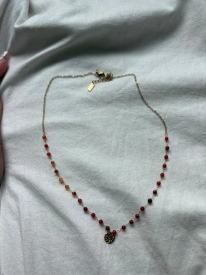 Halsband med röda pärlor och guldberlock - Snyggt halsband med små röda och orange pärlor på tunn guldfärgad kedja. I mitten hänger en rund guldberlock med ett mönster. Perfekt för dig som gillar färg och detaljerade smycken.