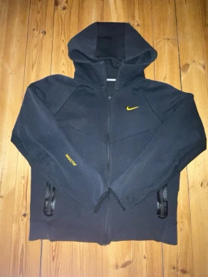 Svart Nike NOCTA hoodie med dragkedja - Säljer en svart hoodie från Nike NOCTA med gul logga på bröstet och NOCTA-tryck på ärmen. Hoodien har dragkedja framtill, två fickor med dragkedja och stor huva. Materialet är mjukt och bekvämt, perfekt för en chill och sportig look.
