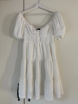 Vit kort klänning från Gina Tricot - Söt vit kort klänning från Gina Tricot med puffärmar och knyt framtill. Klänningen har en smockad överdel, volangdetaljer och är tillverkad i ett lätt, strukturerat tyg. Perfekt för varma dagar och somriga tillfällen.