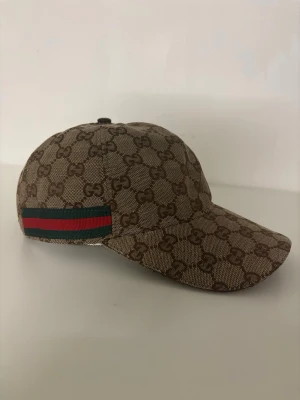 Gucci keps - Säljer en beige keps från Gucci med klassiskt GG-monogram i brunt över hela kepsen. På sidan finns en ikonisk grön och röd rand. Kepsen har böjd skärm och justerbar passform. Materialet är canvas med insida i ljust tyg.