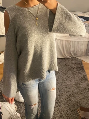 Grå stickad tröja med cold shoulder - Snygg grå stickad tröja med trendiga cold shoulder-detaljer och vida ärmar. Tröjan har rund halsringning och ribbstickad kant nedtill. Perfekt för dig som vill ha en avslappnad men ändå stilren look. Nopprig, därav priset. Borde gå bort med en nopp maskin har inte testat🥰