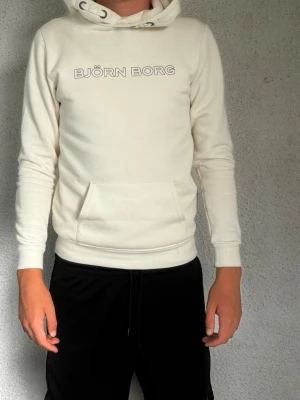 Vit hoodie från Björn Borg - Snygg vit hoodie från Björn Borg med tryckt logga på bröstet. Hoodien har huva med dragsko, magficka och ribbade muddar vid ärmar och nederkant. Tillverkad i mjukt material som känns skönt mot huden. Perfekt för en avslappnad och sportig stil.