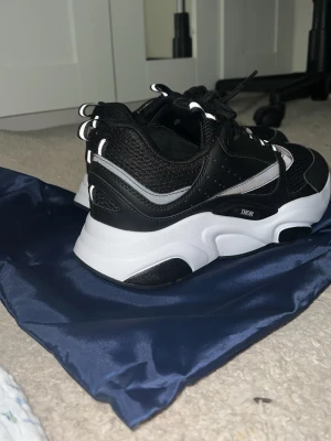 Svarta Dior sneakers med vita detaljer - Chunky sneakers från Dior i svart mesh och läder med vita och grå detaljer. Tjock vit sula och svarta partier undertill. Skorna har svarta snören och reflekterande detaljer på hälen. Perfekta för dig som gillar statement sneakers.