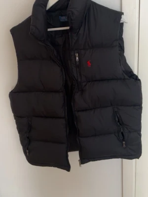 Svart dunväst från Polo Ralph Lauren - Snygg svart dunväst från Polo Ralph Lauren med röd broderad logga på bröstet. Säljer eftersom den va för stor för mig.