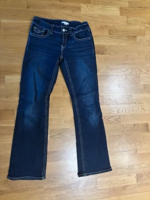 Mörkblå bootcut jeans Gina Tricot - Snygga mörkblå bootcut jeans med vita kontrastsömmar och klassiska fem fickor. Jeansen har lätt slitna detaljer framtill och bakfickor med lock och knapp. Perfekta för en avslappnad och trendig look.