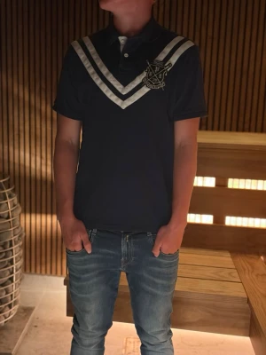 Marinblå pikétröja med vita ränder - Snygg marinblå pikétröja med vita V-formade ränder över bröstet och ett broderat emblem på vänster sida. Tröjan har korta ärmar, krage och knappar framtill. Perfekt för en avslappnad men stilren look.