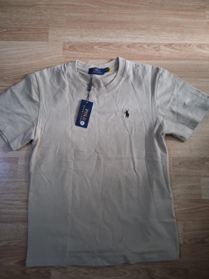 Beige t-shirt från Polo Ralph Lauren - Snygg beige t-shirt från Polo Ralph Lauren med klassisk svart broderad logga på bröstet. T-shirten har rund halsringning och korta ärmar. Tillverkad i mjuk bomull för en skön känsla och enkel stil.