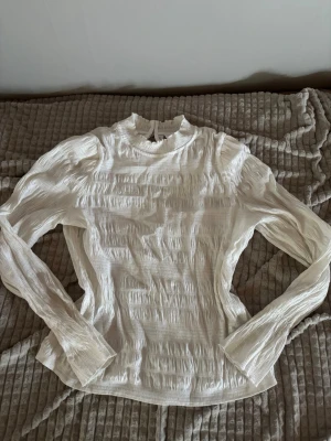Vit plisserad blus med hög krage - Säljer en vit plisserad blus med långärmad design och hög krage. Blusen har fina detaljer med små knappar i nacken och lätt rynkad struktur i tyget. Perfekt för dig som gillar en stilren och trendig look. Skulle s