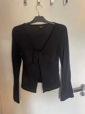 Svart långärmad topp med knyt - Trendig svart långärmad topp med snygg knytning framtill och djup v-ringning. Modellen är croppad och har en smickrande passform som passar perfekt till både jeans och kjol. Tillverkad i mjukt och stretchigt material för extra komfort.