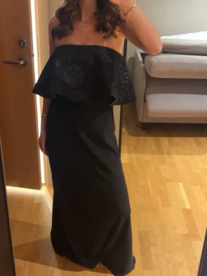 Svart axelbandslös långklänning - Säljer en svart axelbandslös långklänning med volang upptill. Klänningen har en elegant och enkel design med en rak, lång kjol och en snygg offshoulder-look. Perfekt för dig som vill ha en stilren och trendig outfit.