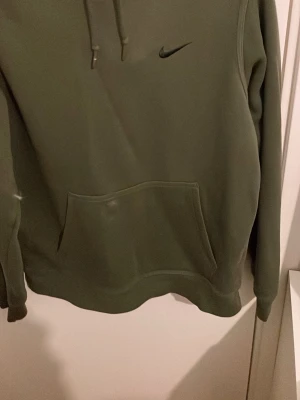 Olivgrön hoodie från Nike - Säljer en olivgrön hoodie från Nike med klassisk huva och dragsnören. Hoodien har en stor magficka framtill och Nike-logga broderad på bröstet. Tillverkad i mjukt material som passar perfekt för en avslappnad stil.