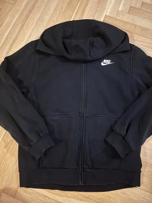 Svart zip hoodie från Nike - Säljer en svart hoodie från Nike med dragkedja framtill och vit Nike-logga på bröstet. Hoodien har huva, ribbade muddar vid ärmar och nederkant samt en klassisk streetwear-look. Perfekt för dig som gillar enkel och sportig stil.
