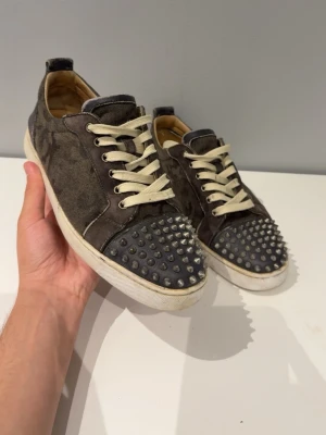 Christian Louboutin  - Säljer ett par mörkgrå sneakers med coola silvriga nitar på tån och vita snören. Skorna har en låg profil och är gjorda i mockaliknande material med vit sula. Perfekta för dig som vill sticka ut med din stil.