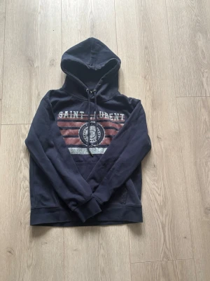 Mörkblå Saint Laurent hoodie - Mörkblå Hoodie från Saint Laurent använd ca 5 gånger så nästan helt ny. Sitter bra i längden på mig men kort i armarna jag är 176 och väger 56.
