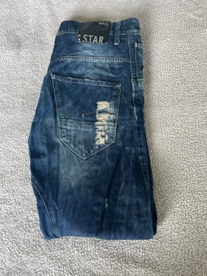 Blå  jeans från G-Star med slitningar - Säljer ett par blå jeans från G-Star med slitningar och tvättade detaljer. Jeansen har raka ben, klassiska bakfickor och en cool, avslappnad look. Perfekta för dig som gillar streetwear och en lite tuffare stil.