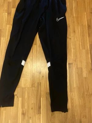 Svarta Nike träningsbyxor  - Säljer ett par svarta träningsbyxor från Nike med vita detaljer vid knäna och klassisk  vit Nike-logga på låret. Byxorna har resår i midjan och vid bensluten, samt en avslappnad passform. Perfekta för chill eller träning.