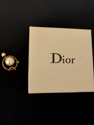 Dior örhänge med pärldetaljer - Unikt örhänge från Dior i guldfärgad metall med två vita pärlor. Designen har en cirkel med en liten stjärna och måne, vilket ger en cool och elegant vibe. Super rare vintage.