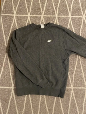 Mörkgrå sweatshirt från Nike - Snygg mörkgrå sweatshirt från Nike med klassisk vit logga broderad på bröstet. Tröjan har rund halsringning, långa ärmar och ribbade muddar vid ärmslut och nederkant. Perfekt för en avslappnad och sportig stil.