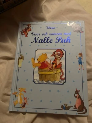 Visor och ramsor med Nalle Puh - En ljusblå barnbok med hårda pärmar, fylld med visor och ramsor från Nalle Puhs värld. Omslaget pryds av Nalle Puh, Tiger och flera andra karaktärer från Sjumilaskogen. Boken är rikt illustrerad med färgglada bilder och passar perfekt för högläsning.