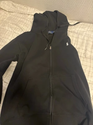Svart hoodie från Polo Ralph Lauren - Säljer en svart hoodie från Polo Ralph Lauren med dragkedja, huva med dragsko och två stora fickor framtill. Klassisk vit logga broderad på bröstet. Perfekt för en chill och stilren look. Storlek L men passar för S 