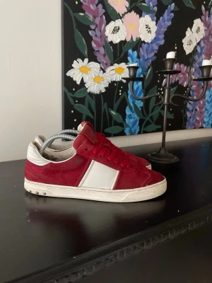 Valentino Flycrew  - Snygga röda sneakers från Valentino med vita detaljer på sidan och vit sula. Skorna har en klassisk låg modell och är gjorda i mocka och läder. Perfekta för dig som vill sticka ut med en stilren och trendig look.