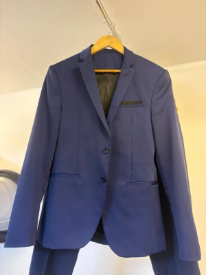 Blå kostym med tvåknäppt kavaj - Snygg blå kostym med klassisk tvåknäppt kavaj och raka byxor. Kavajen har smal passform, svarta detaljer vid bröstfickan och två fickor framtill. Perfekt för dig som vill ha en stilren och modern look.